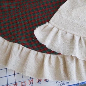 May include: A red and green plaid fabric with a white ruffled trim. The trim is folded over and sewn to create a double layer of ruffles.