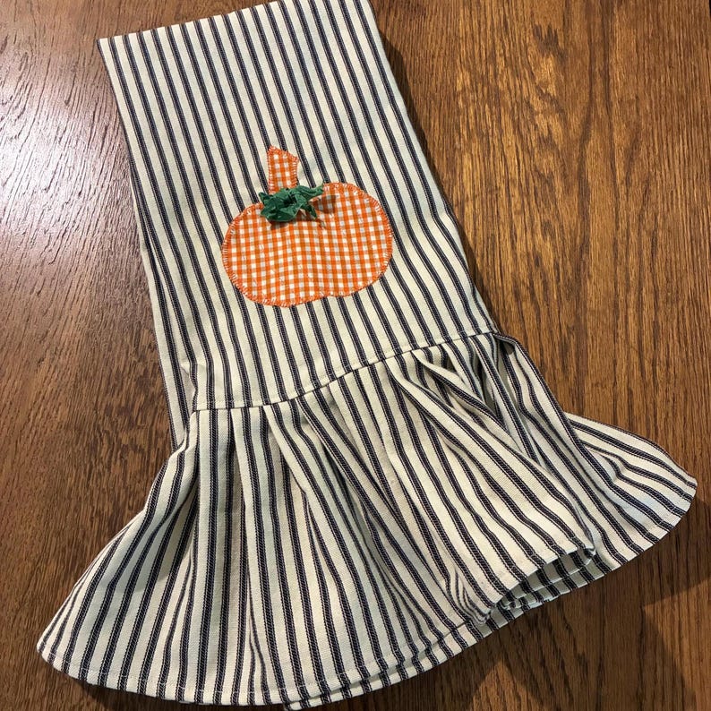Op de afbeelding: Een decoratieve keukendoek met een blauw-wit streepjespatroon. Een oranje en witte gingham pompoen applicatie is op de bovenkant genaaid. De onderkant van de doek heeft een gefranjerde rand.