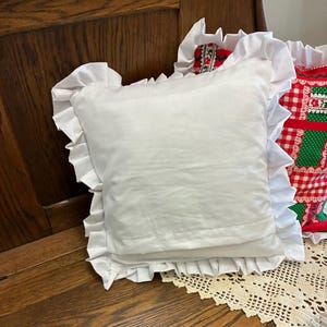 May include: A square white throw pillow featuring a ruffled border along all four edges. The pillow is made of a solid white fabric and is positioned in front of another pillow that has a red, white, and green checkered pattern with floral details.