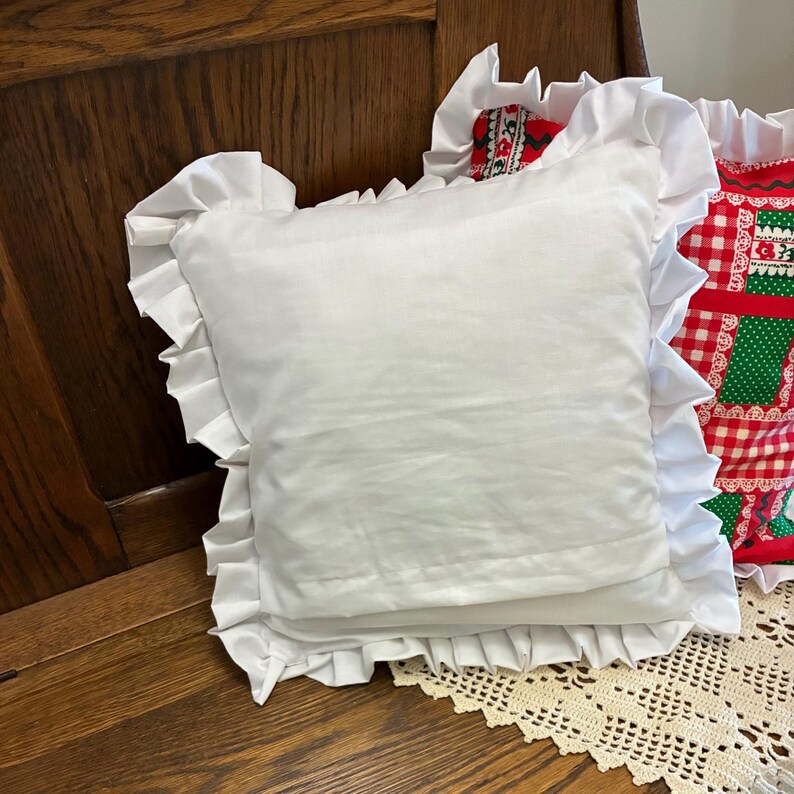 May include: A square white throw pillow featuring a ruffled border along all four edges. The pillow is made of a solid white fabric and is positioned in front of another pillow that has a red, white, and green checkered pattern with floral details.
