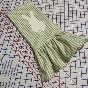 Op de afbeelding: Een gestreepte, cr&egrave;mekleurige en lichtgroene keukendoek met Pasen als thema. De handdoek heeft een witte konijnenapplicatie en een gefranjerde rand. De stof heeft verticale strepen. De handdoek is gevouwen en schuin op een patroon.
