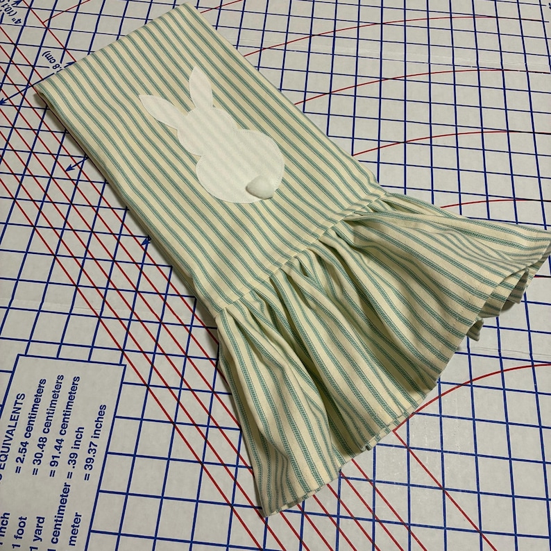 Op de afbeelding: Een gestreepte, cr&egrave;mekleurige en lichtgroene keukendoek met Pasen als thema. De handdoek heeft een witte konijnenapplicatie en een gefranjerde rand. De stof heeft verticale strepen. De handdoek is gevouwen en schuin op een patroon.