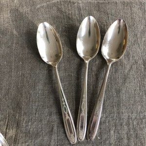 May include: Three silver spoons with ornate designs on a gray fabric background.