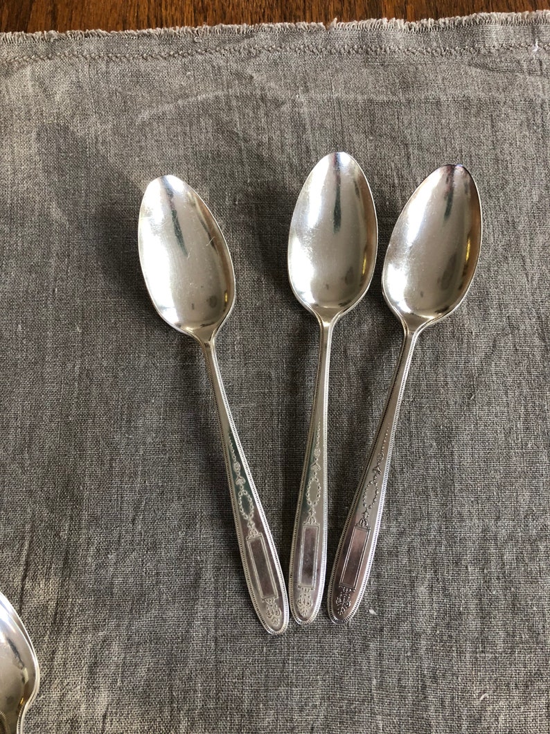 May include: Three silver spoons with ornate designs on a gray fabric background.