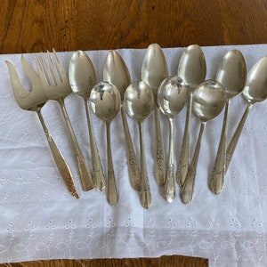 May include: A set of 10 silver spoons and two silver forks. The spoons have ornate designs and are arranged on a white cloth background.