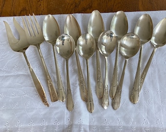 May include: A set of 10 silver spoons and two silver forks. The spoons have ornate designs and are arranged on a white cloth background.