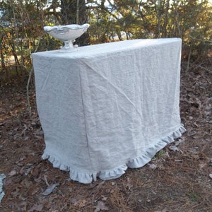 May include: A rectangular table covered in a beige linen cloth with a ruffled hem, featuring a white decorative pedestal bowl with silver-toned accents placed on the top surface. The fabric has a visible weave and drapes over the sides.