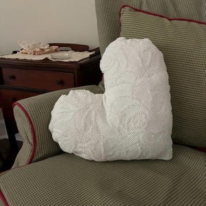 May include: A white, heart-shaped decorative pillow with a textured surface and a floral pattern. The pillow is placed on a patterned armchair with red trim. A small wooden table is visible in the background.