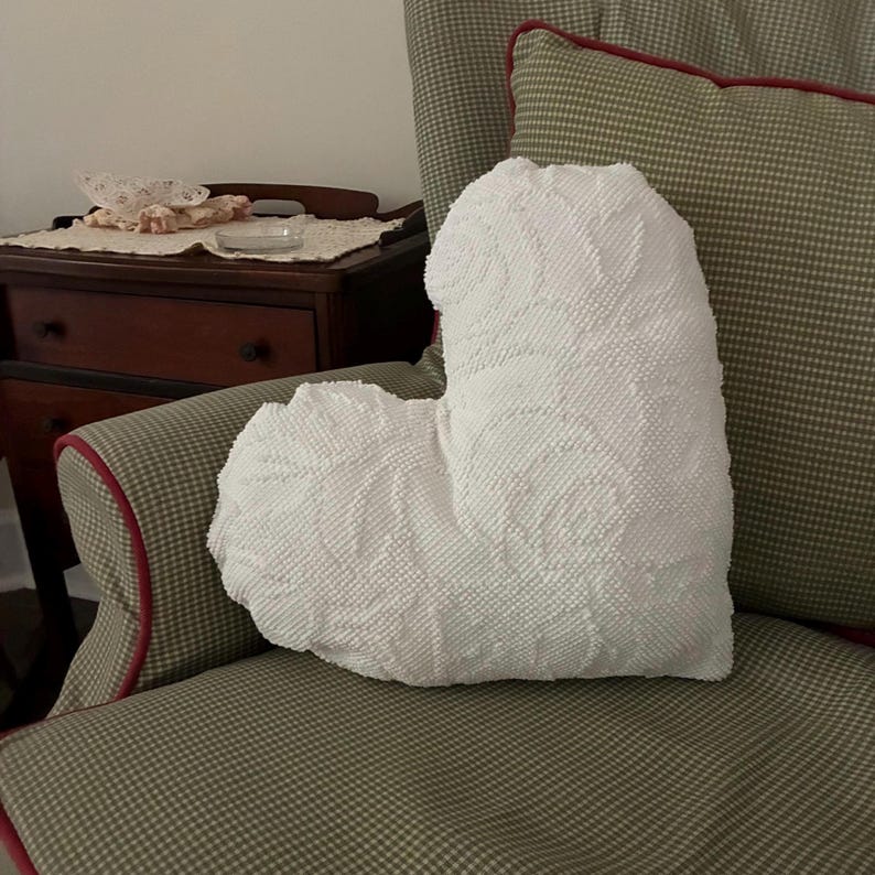 May include: A white, heart-shaped decorative pillow with a textured surface and a floral pattern. The pillow is placed on a patterned armchair with red trim. A small wooden table is visible in the background.