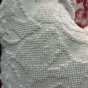 May include: Close-up of a white, textured fabric with a raised, tufted pattern. The fabric has a soft, plush appearance, with a repeating geometric design. The texture is reminiscent of a towel or blanket.