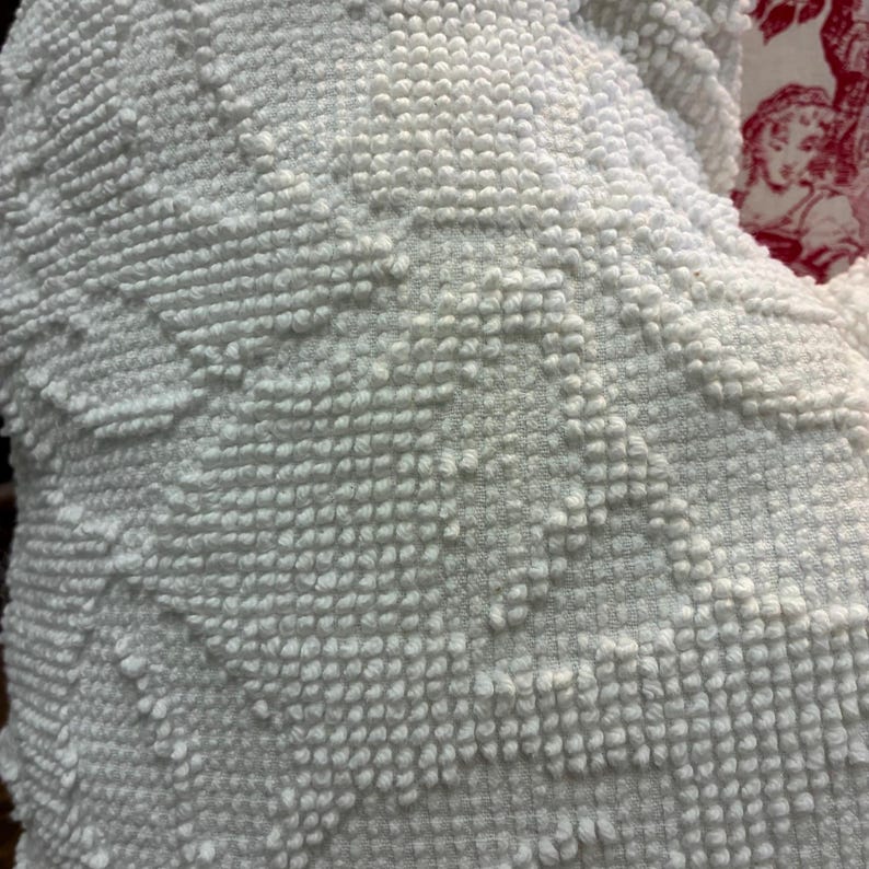 May include: Close-up of a white, textured fabric with a raised, tufted pattern. The fabric has a soft, plush appearance, with a repeating geometric design. The texture is reminiscent of a towel or blanket.