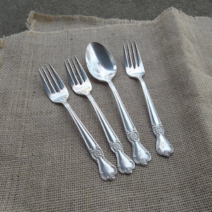 May include: Four pieces of silverware, including a spoon and three forks, are arranged on a brown burlap surface. The silverware is silver in color and has an ornate design. Each piece has a letter "G" engraved on the handle.