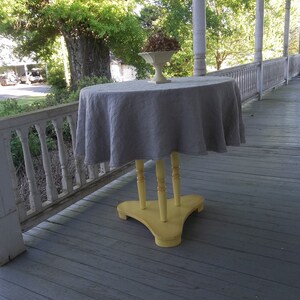 May include: A gray linen tablecloth draped over a yellow wooden table with three legs. The table is on a wooden porch with white railings.