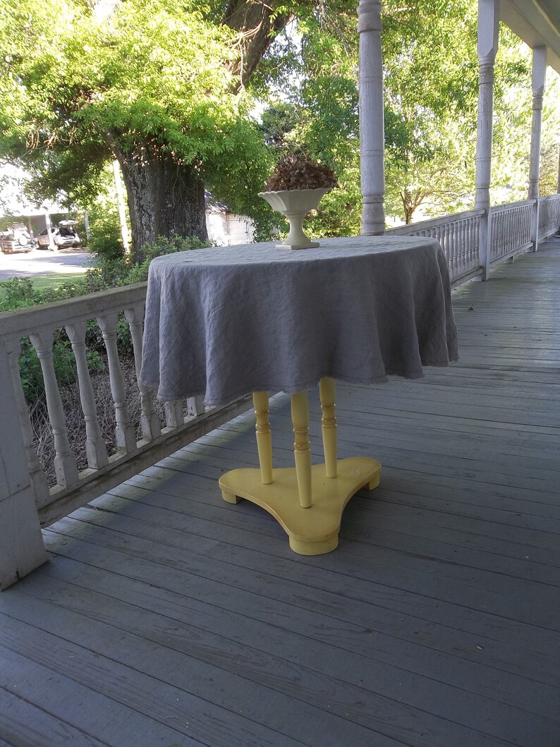 May include: A gray linen tablecloth draped over a yellow wooden table with three legs. The table is on a wooden porch with white railings.