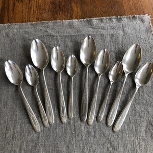 May include: A set of ten silver spoons with ornate designs. The spoons are arranged in a row on a gray fabric surface.