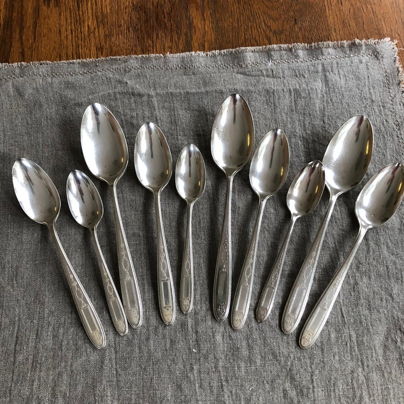 May include: A set of ten silver spoons with ornate designs. The spoons are arranged in a row on a gray fabric surface.