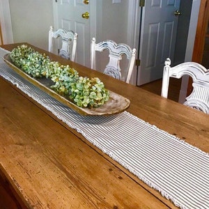 May include: A wooden table runner with a white and black striped pattern and a white crocheted edge. A wooden dough bowl with green hydrangeas sits on top of the runner.