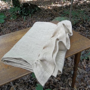 May include: A natural burlap table runner with a ruffled edge. The runner is folded and placed on a wooden table. The table is outdoors on a bed of brown leaves.