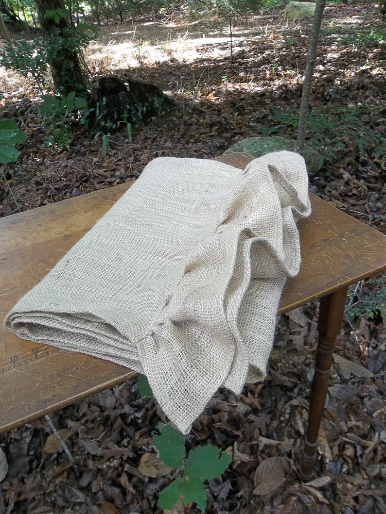 May include: A natural burlap table runner with a ruffled edge. The runner is folded and placed on a wooden table. The table is outdoors on a bed of brown leaves.