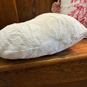 May include: A white, textured pillow with a rounded shape. The pillow is made of small, tightly packed, raised elements, creating a bumpy surface. The pillow is resting on a wooden surface.