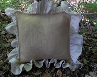 May include: A beige burlap pillow with a white ruffled trim. The pillow is lying on a bed of brown leaves.