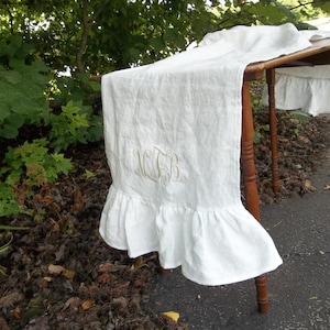 May include: White linen table runner with a ruffled edge and embroidered monogram. The runner is draped over a wooden table, showcasing its texture and design. The monogram is in a light yellow color.