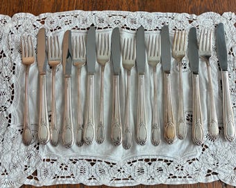 May include: A collection of fourteen silver-toned metal flatware pieces, including seven dinner knives and seven forks, arranged in an alternating pattern on a white rectangular lace doily with intricate floral embroidery.