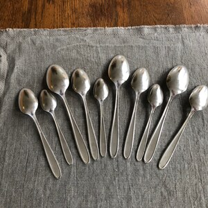 May include: A collection of eleven silver spoons arranged in a semi-circle on a gray fabric surface.