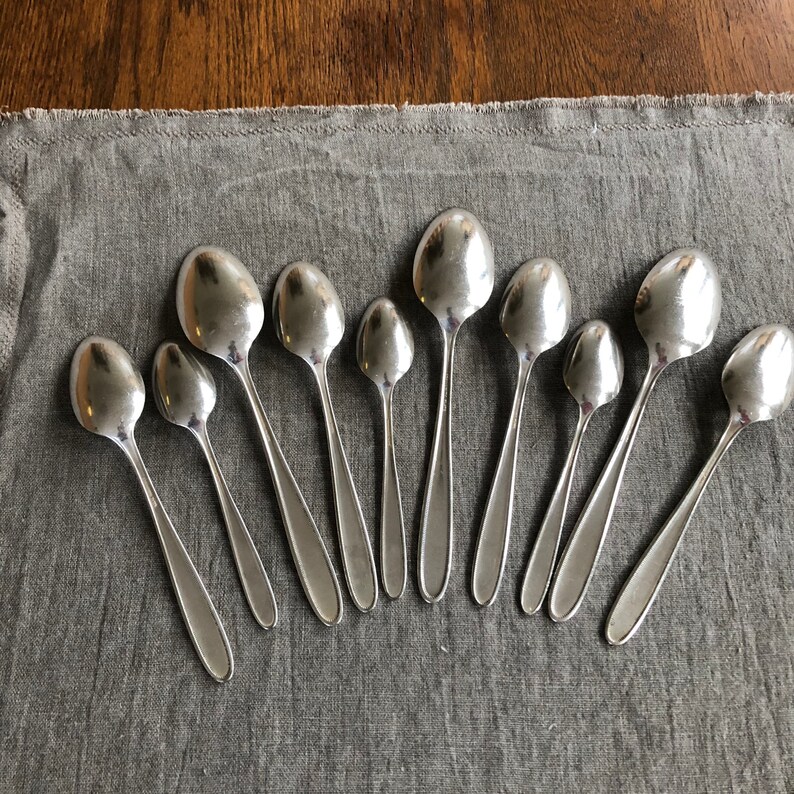 May include: A collection of eleven silver spoons arranged in a semi-circle on a gray fabric surface.