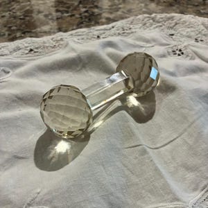 May include: A clear crystal dumbbell-shaped object with faceted spherical ends and a rectangular center. The crystal reflects light, creating a sparkling effect. The object is resting on a white, textured surface.