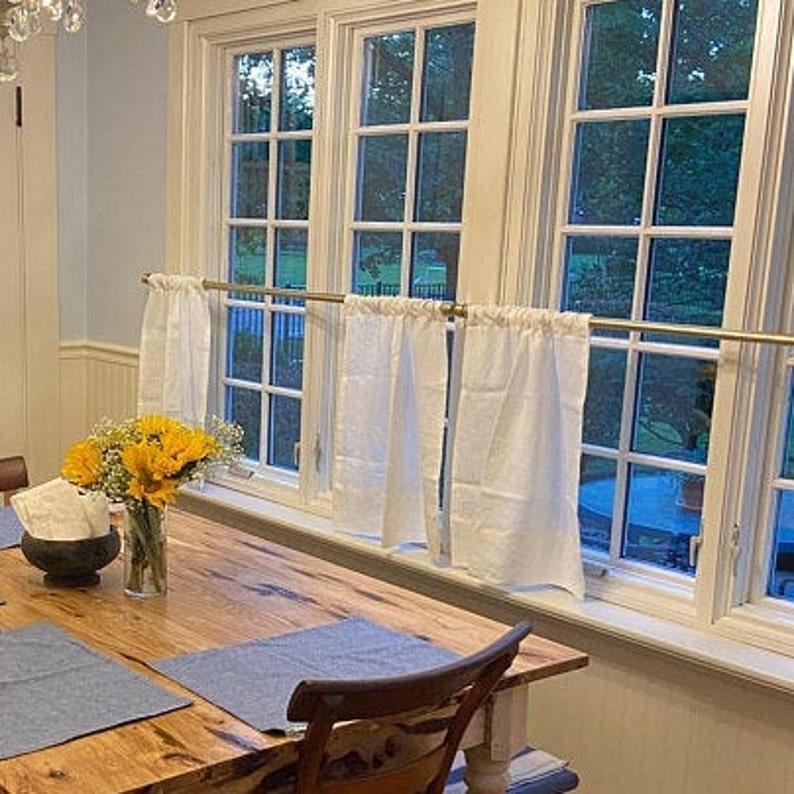May include: A wooden dining table with a blue placemat and a chair. The table is set with a vase of yellow flowers. There are white curtains hanging in the window behind the table.