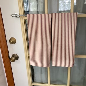 Op de afbeelding: Twee keukendoeken met verticale bordeauxrode en cr&egrave;mekleurige strepen hangen aan een zilveren stang. De doeken zijn gemaakt van een zachte stof en worden gepresenteerd op een glazen deur. De deur heeft een messing deurknop en slot.