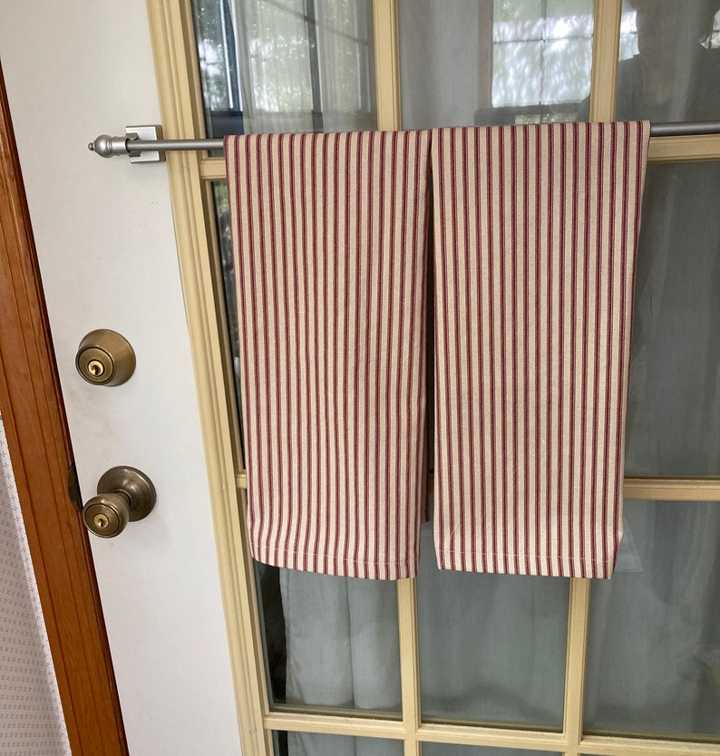 Op de afbeelding: Twee keukendoeken met verticale bordeauxrode en cr&egrave;mekleurige strepen hangen aan een zilveren stang. De doeken zijn gemaakt van een zachte stof en worden gepresenteerd op een glazen deur. De deur heeft een messing deurknop en slot.