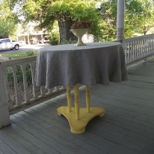 May include: A gray linen tablecloth draped over a yellow painted table with three legs. The table is on a wooden porch with a white railing.