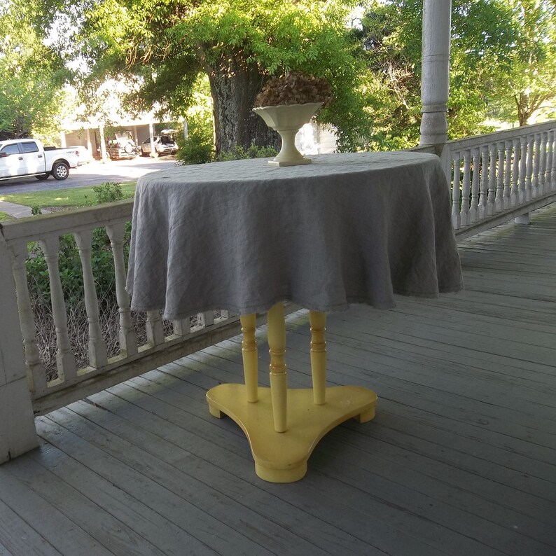 May include: A gray linen tablecloth draped over a yellow painted table with three legs. The table is on a wooden porch with a white railing.