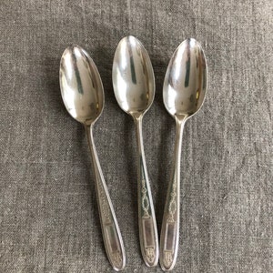 May include: Three silver teaspoons with ornate handles. The spoons are arranged in a row on a textured, light brown fabric background.