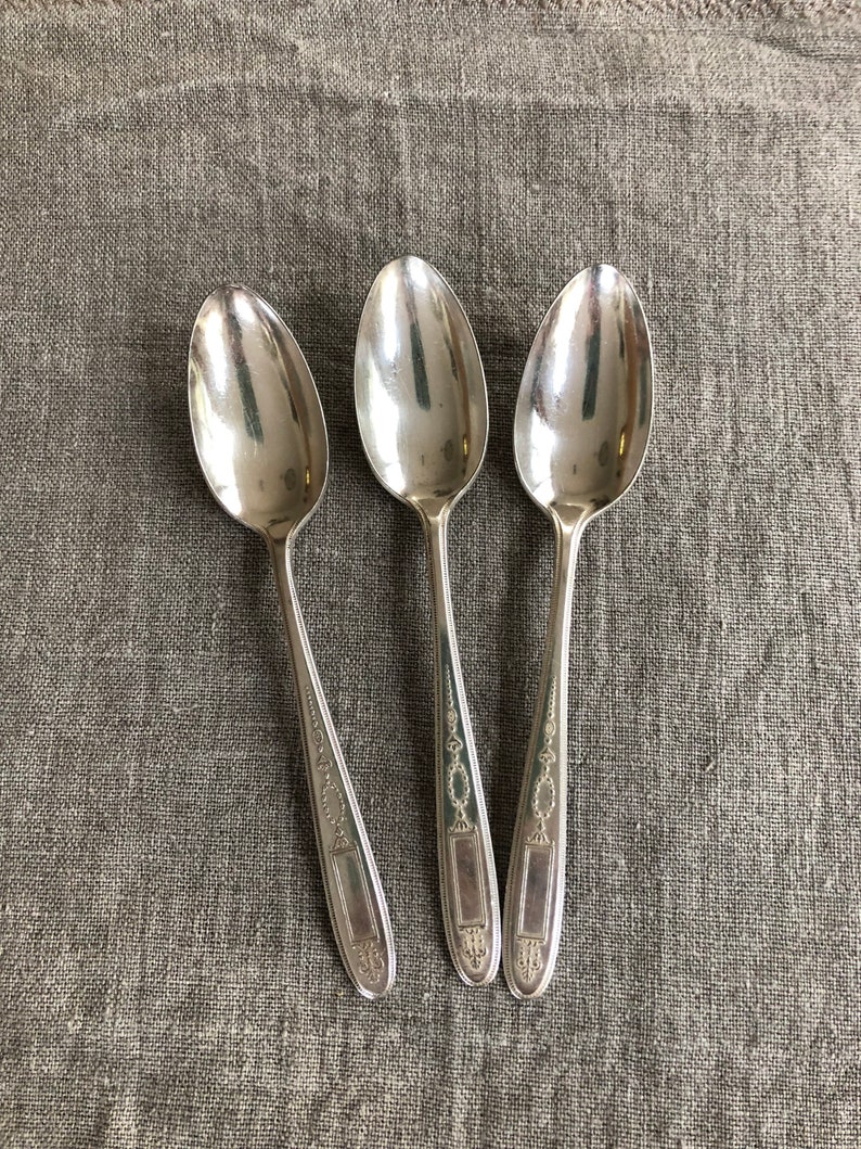 May include: Three silver teaspoons with ornate handles. The spoons are arranged in a row on a textured, light brown fabric background.