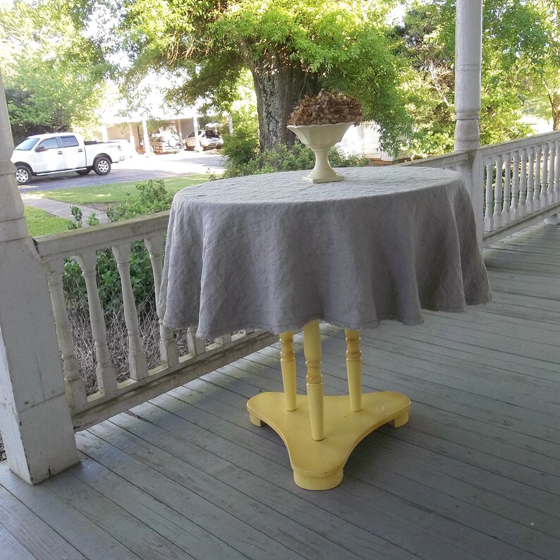May include: A gray linen tablecloth draped over a yellow wooden table with a white pedestal bowl on top. The table is on a porch with a white railing.