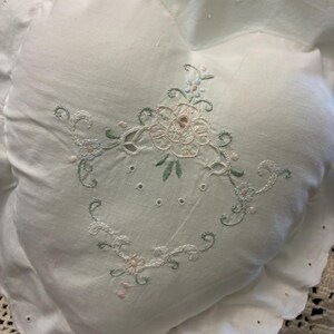 May include: A heart-shaped white pillow featuring delicate floral embroidery in soft pink and sage green thread. The pillow has a scalloped edge with eyelet detailing around the perimeter, creating a decorative border on the white fabric surface.