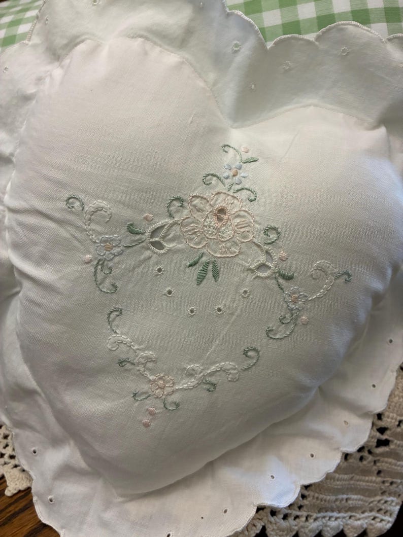 May include: A heart-shaped white pillow featuring delicate floral embroidery in soft pink and sage green thread. The pillow has a scalloped edge with eyelet detailing around the perimeter, creating a decorative border on the white fabric surface.
