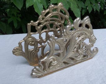 May include: Ornate, gold-colored metal napkin holder with a decorative, openwork design. The holder features scrolling floral and leaf patterns. The napkin holder is displayed on a white surface with a green leafy background.