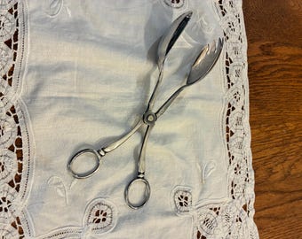 May include: A pair of silver-toned metal serving tongs with two ring-shaped handles and a central pivot point. One side features a solid, smooth, rounded spoon-like bowl, while the other side has a matching bowl with three distinct, pointed tines.