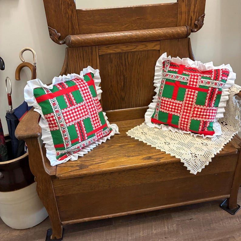 May include: Two square throw pillows featuring a patchwork pattern of red and white gingham, green polka dots, and floral strips. Each pillow is finished with a white ruffled border and is placed on a wooden bench with a cream lace doily.