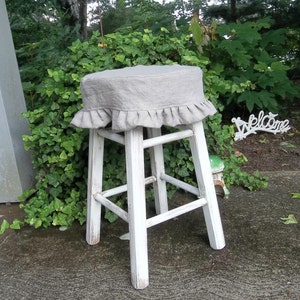 May include: White wooden stool with a gray linen seat cover with a ruffled edge. The stool is sitting on a concrete patio.