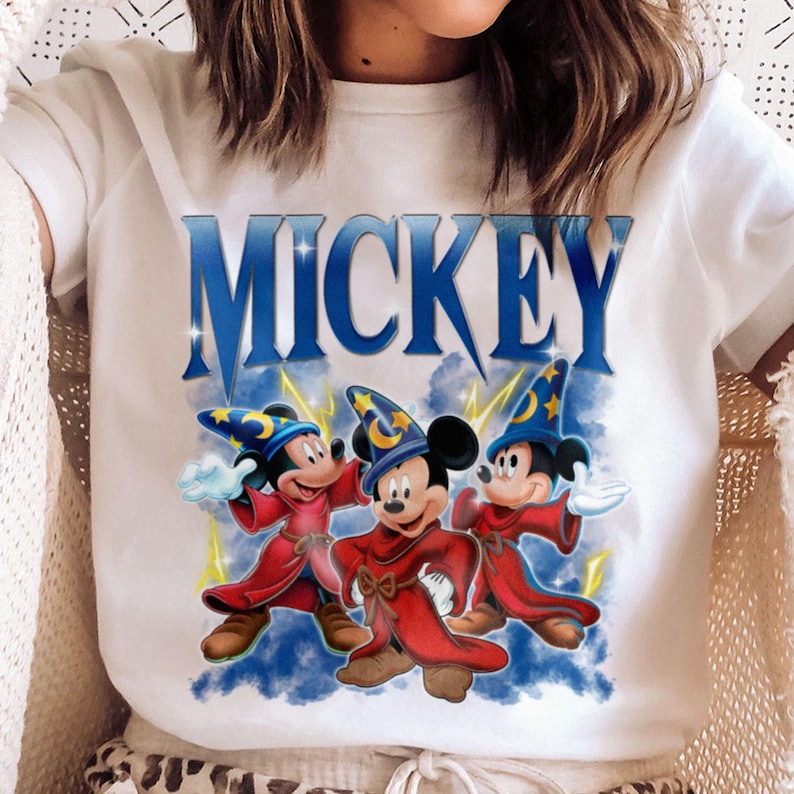 Puede incluir: Camiseta blanca con la palabra "MICKEY" en azul, sobre un gr&aacute;fico de tres personajes de Mickey Mouse con t&uacute;nicas rojas y sombreros de mago. El dise&ntilde;o incluye rayos amarillos y un fondo de nubes azules.