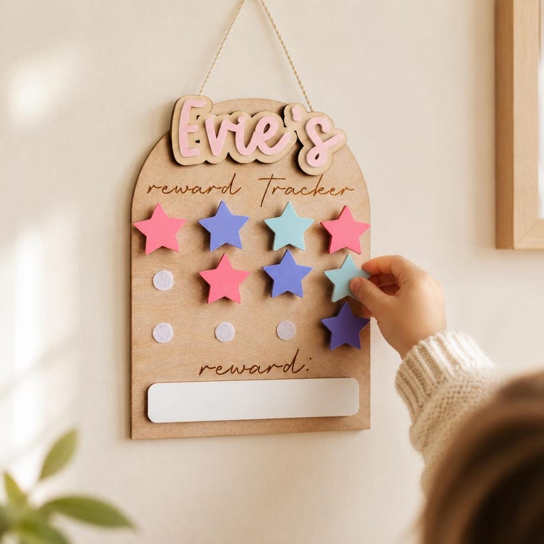 Może przedstawiać: Drewniana tablica nagr&oacute;d z imieniem "Evie's" w r&oacute;żowych literach. Tablica posiada magnetyczne gwiazdki w kolorze r&oacute;żowym, niebieskim i turkusowym. Słowa "reward tracker" i "reward" są napisane na tablicy. Ręka umieszcza gwiazdkę na tablicy.