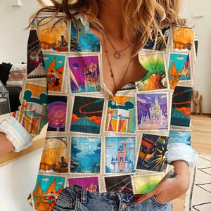 May include: A long-sleeved shirt featuring a collage of colorful travel-themed postage stamp designs. The shirt has a button-down front and is paired with blue denim jeans. The stamps depict various landmarks and attractions.