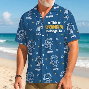 May include: Blue button-down shirt with a repeating pattern of cartoon children, stars, and names. The text "This Grandpa Belongs To" is in the center. The shirt has short sleeves and is worn on a beach.