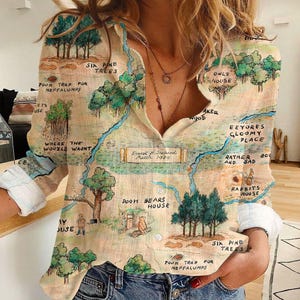 May include: Beige button-down shirt featuring a map-like illustration with text and drawings of trees, houses, and characters. The shirt has long sleeves with white cuffs and a classic collar. The map includes text such as "Pooh Bears House" and "Six Pine Trees."