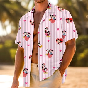 May include: A short-sleeved pink button-down shirt with white vertical stripes. The shirt features a pattern of Mickey and Minnie Mouse in heart-shaped designs, along with pink hearts. The words "Happy Valentine's Day" are visible on the hearts.